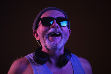 Fototapeta na wymiar a cool aged man in hipster hat, sunglasses and tank top listening to music via headphones and singing along, dancing. Isolate on dark background.
