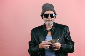 Stock footage of a senior hipster man in leather jacket, grey hat, sunglasses and earpods throwing dollar banknotes from the stack. Isolate on pink background.