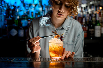 Woman bartender decorates glass with cocktail by smoking stick