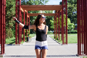 Young pretty brunette woman, riding rollerblades in city park with green trees. Fit sporty girl, wearing black top and white shorts, roller-skating, raising her arms up, showing joy and happiness.