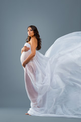 Studio portrait of nude pregnant woman in a white flying dress fabric on grey background. Beautiful healthy pregnancy, happy motherhood