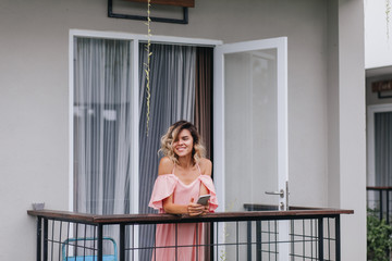 Winsome tanned girl with phone in hands smiling and looking away. Cheerful young lady in pink outfit standing at hotel balcony.