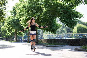 Young pretty brunette woman, riding rollerblades in city park with green trees. Fit sporty girl, wearing protective outfit, roller-skating in summer. Full-length portrait of slim sportswoman.