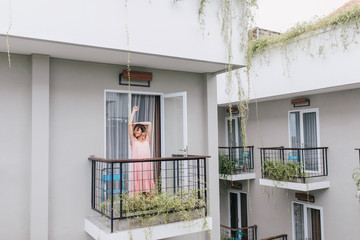 Joyful girl with blonde hair enjoying morning at terrace. Photo of smiling curly female model stretching at hotel balcony.