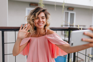 Cheerful tanned girl using smartphone for selfie at balcony. Magnificent blonde lady in pink attire taking picture of herself and waving hand.