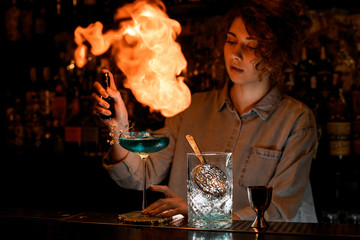 Young woman at dark bar professionally makes fire over wineglass with cocktail.