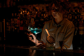 Young woman at dark bar begin to makes fire over wineglass with blue drink.