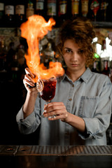 Professional woman barman holds wineglass with cocktail and makes flame over it.
