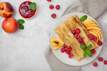 Crepes, thin pancakes with raspberries and peaches, mint and jam on a plate. Light background. Top view. Flat lay. Copy space.