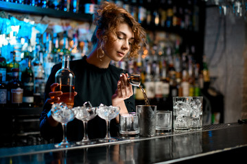 Young barman lady carefully pours drink into metal glass using jigger - obrazy, fototapety, plakaty