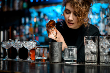 Young pretty woman at bar pours drink into metal glass using steel jigger.