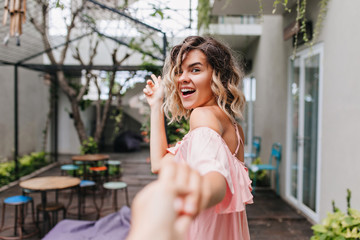 Fototapeta premium Excited girl with wavy hair walking near outdoor cafe. Pleased blonde woman with tanned skin going to street restaurant.