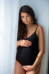 A beautiful pregnant young woman in a black body suit is standing near a window with curtains. Sunny day, white light home interior