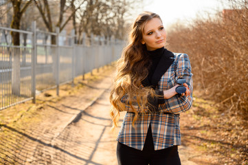 Portrait of a stylish beautiful young girl with long hair with a smile in the park on the background of the road. Photo taken in sunny weather outdoors in spring.