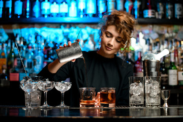 Young woman barman preparing cocktail and carefully pouring it into glass. - obrazy, fototapety, plakaty