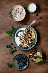 Homemade crunchy puffed millet grain granola with dried fruits and nuts in ceramic bowl, with yogurt, mint and ingredients above. Brown texture background. Flat lay, space. Healthy food breakfast