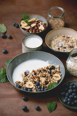Homemade crunchy puffed millet grain granola with dried fruits and nuts in ceramic bowl, with yogurt, mint and ingredients above. Brown texture background. Close up. Healthy food breakfast