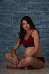 Fototapeta na wymiar Beautiful pregnant young athletic woman in sports maternity underwear is sitting against the background of a gray brick wall