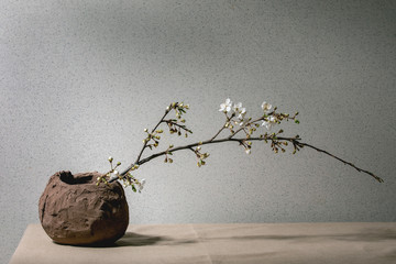Blossom cherry branches in craft clay vase on grey table cloth. Spring interior decorations.