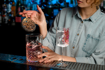 Young woman barman holds in her hand strainer over large glass with pink drink