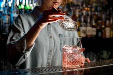 Close-up young woman's hand holds strainer over large glass with pink drink