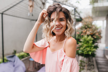 Close-up portrait of adorable young woman posing with surprised smile in street restaurant. Tanned girl in pink blouse playing with her light wavy hair.