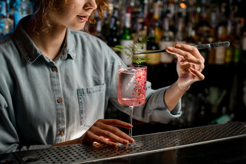 young woman barman decorates glass with rose cocktail by piece of green plant.