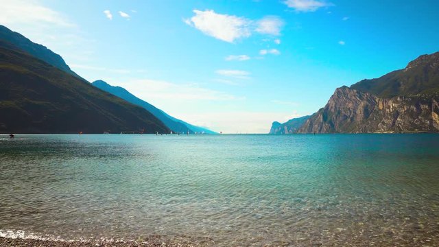 Panoramic views of Lake Garda in resort Torbole Nago. Azure sky clouds Italian alpine alps. Clear water surf wind waves. Tourists enjoy windsurfing surfing sports activities. Lago di Garda Benaco.