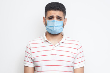 Portrait of sad upset or bored man with surgical medical mask in striped t-shirt standing and looking at camera with dissatisfied sadness face. indoor studio shot, isolated on white background.