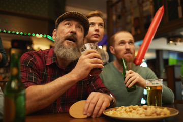 Mature bearded man in cap holding beer while sitting at the table with his friends and they watching sport game in the bar