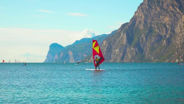Windsurfer red sailing floats on water surface of Lake Garda in Torbole Nago resort. Rowing, sports. Backdrop group tourists, sky clouds foggy haze wind, Italian Alpine mountains. Lago di Garda Benaco