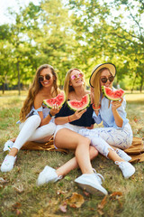 Three beautiful young girls have fun together and eating watermelon  in hot summer day. Friends holding slice of watermelon and posing in the park. Summer concept. 