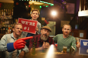 Mature men and woman with placard sitting at the table with beer and cheering for their football team in sport bar