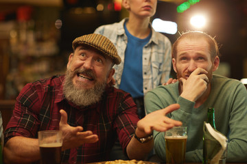 Mature men gesturing and looking unsatisfied while watching football match and drinking beer in sport bar