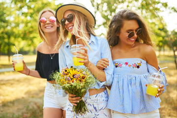 Fototapeta premium Three beautiful young girls have fun together and drink fresh fruit juice in hot summer day. Friends enjoying orange juice and posing against the backdrop of the park. Summer concept.