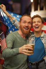 Fototapeta premium Happy young couple drinking beer and cheering for the American football team in sport bar