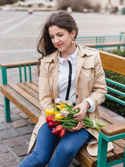 Beautiful young woman with spring tulips flowers bouquet at city street. Happy girl sitting on a bench outdoors. Spring portrait of pretty female in old town