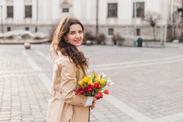 Fototapeta premium Beautiful young woman with spring tulips flowers bouquet at city street. Happy girl walking outdoors. Spring portrait of pretty female in old town