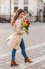 Fototapeta premium Beautiful young woman with spring tulips flowers bouquet at city street. Happy girl walking outdoors. Spring portrait of pretty female in old town