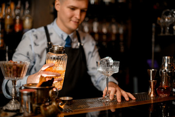 Young barman holds glassy cocktail shaker. Glass with ice stands on bar counter.