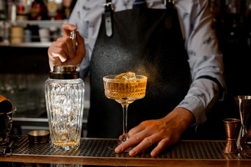 Barman in black apron sprinkles on glass with cocktail.