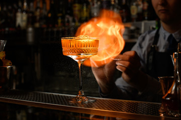 Glass of alcoholic drink and ice stands on bar counter. Bartender makes fire flame behind it.