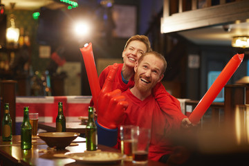Happy young couple of fans in red shirts watching football match and cheering for their team in sport bar