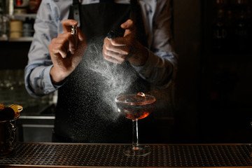 Bartender in black apron sprinkles on glass with cocktail.