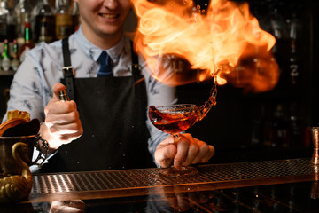 young smiling bartender holds glass with cocktail, spraying on it and sets on fire.