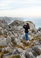 Obraz premium Beautiful young girl with black sweater standing on rocks beside cliffs at the coast in a beautiful landscape