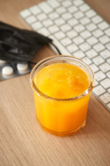 Glass of fresh orange juice on the table in home office. Keyboard, mask and pills are on the table.