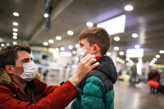 Father Putting Medical Mask On His Son To Protect Himself From The Coronavirus In An Airport Terminal Or Shopping Mall