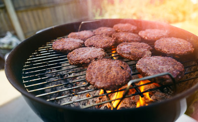 Burgers being cooked on a barbecue grill with flames coming off the charcoal on a sunny day in the garden.