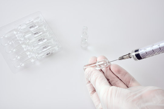 Ampoules with medicine in a blister next to upright ampoule with medicine on white acrylic table, hands in white medical gloves drawing medicine into a syringe
